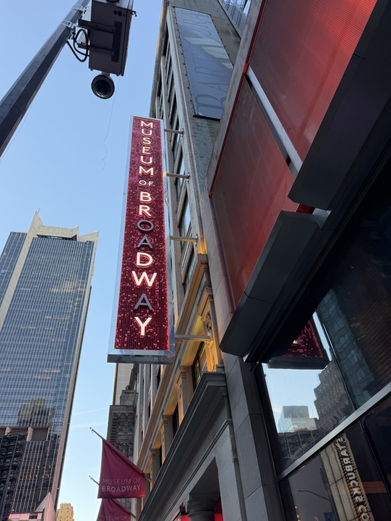View of the Museum of Broadway sign from below, showcasing its red and white lights against a city backdrop.