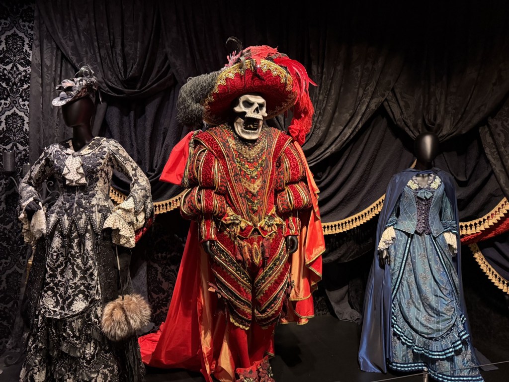 Three mannequins displaying elaborate costumes: a skeleton in a vibrant red and gold outfit, a black and white ruffled dress, and a blue vintage gown.
