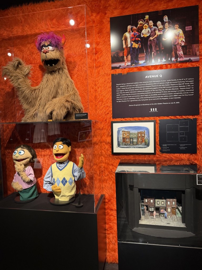 Display of Avenue Q puppets and set design at an exhibition, featuring a furry puppet with purple hair, two additional puppets, and a miniature set model. Background includes a framed photo of the cast and a description of the musical.