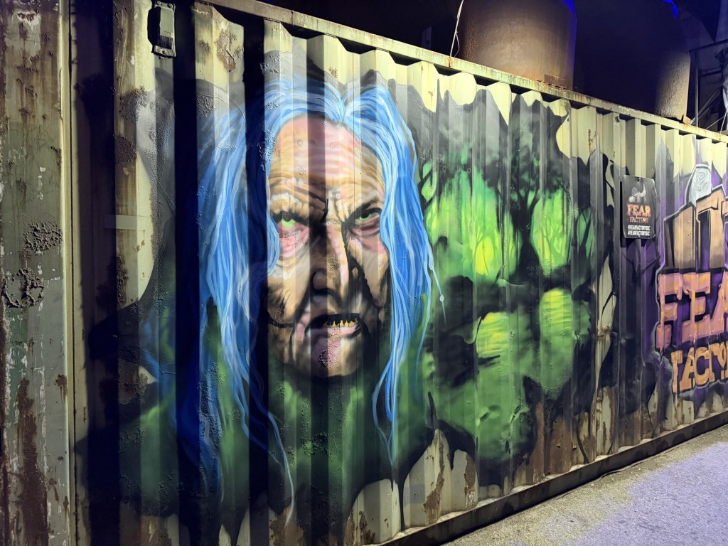 A colorful graffiti mural on a shipping container depicting a sinister-looking elderly woman with long blue hair and an intense expression, surrounded by green hues.