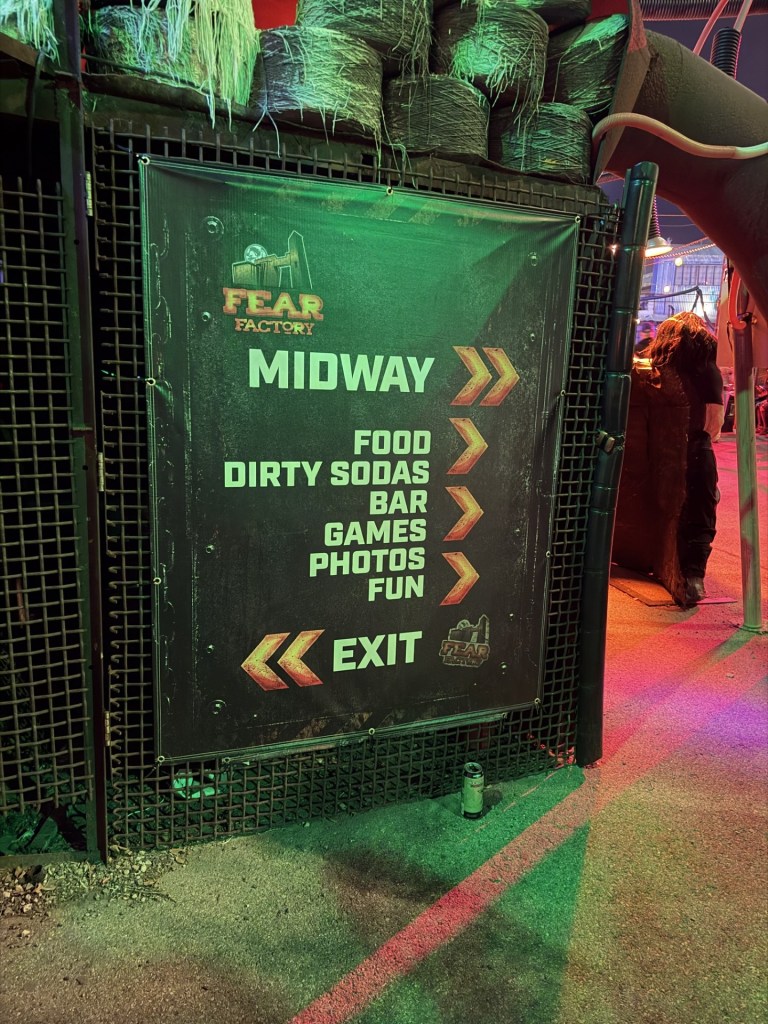 Signage for 'Fear Factory' featuring activities available at the Midway, including food, dirty sodas, bar, games, photos, and fun, along with an exit direction.