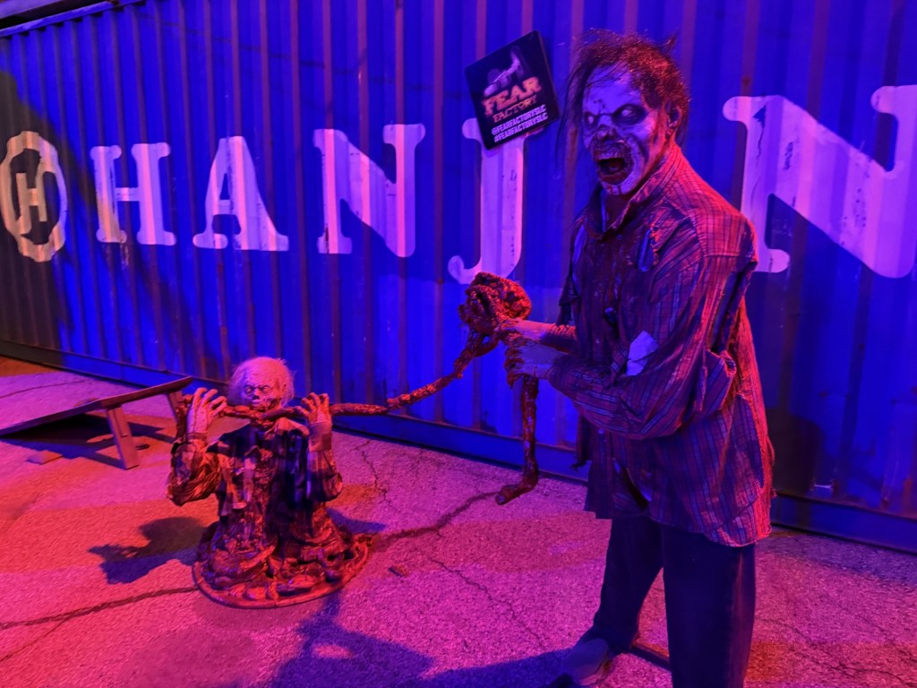 A creepy scene featuring two zombie-like figures. One zombie is standing and holding a rope, while the other is seated, appearing to be in distress. The background includes a shipping container with purple lighting, adding to the eerie atmosphere.