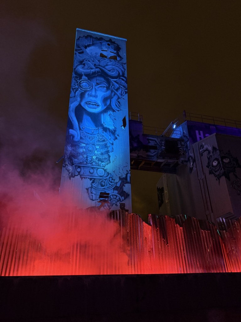 A tall structure with vibrant street art featuring a woman's face, surrounded by mist and illuminated with blue and red lights.