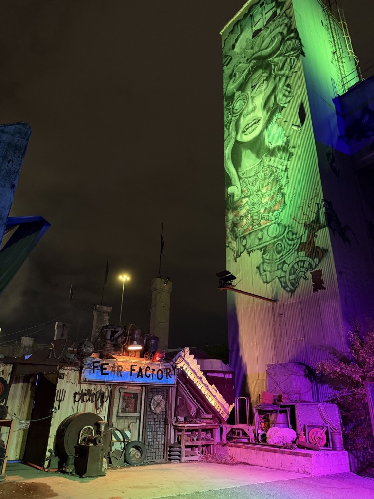An eerie night scene featuring a building labeled 'Fear Factory' with industrial decor, illuminated by colorful lights. A large mural of a face with mechanical elements is painted on the adjacent tower, enhancing the haunting atmosphere.