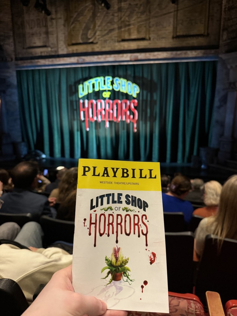 A hand holding a Playbill for 'Little Shop of Horrors' in a theater with a curtain in the background.