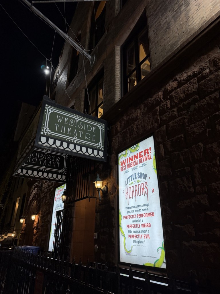 Exterior of the Westside Theatre at night, featuring a marquee for the musical 'Little Shop of Horrors' with details of its award for Best Musical Revival.