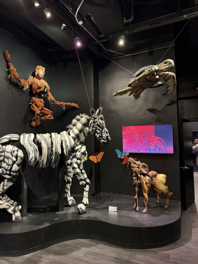 An art installation featuring animal sculptures made of various materials, including a zebra, a lion, a monkey, and a turtle, displayed against a black wall with colorful butterflies and a vibrant artwork in the background.